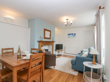 A dining table with chairs and a wine bottle next to a blue sofa and a wooden fireplace in a living room at Spring Cottage in Birdlip near Brockworth