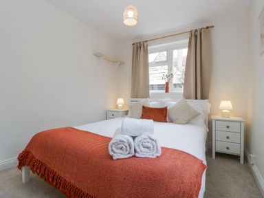A bedroom with a bed with white and orange bedding two bedside tables with lamps and a window with beige curtains at Spring Cottage in Birdlip near Brockworth