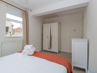A bedroom with a bed covered in white linens and an orange throw with rolled towels on top a white wardrobe with mirrored doors a white chest of drawers and a window with beige curtains at Spring Cottage in Birdlip near Brockworth