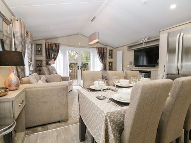 A dining table with tableware and beige chairs next to a living area with sofas and large glass doors at Water's Reach White Cross Bay Holiday Park near Troutbeck Bridge