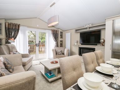 A living room with beige sofas a coffee table dining chairs with place settings a television and glass doors to a balcony at Water's Reach White Cross Bay Holiday Park near Troutbeck Bridge