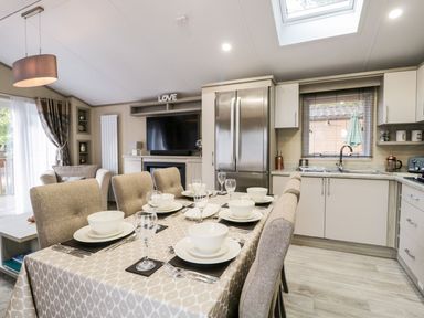 A dining table set with bowls, plates, cutlery and glasses in a kitchen and living room area with a refrigerator, sink, TV and armchair at Waters Reach White Cross Bay Holiday Park near Troutbeck Bridge