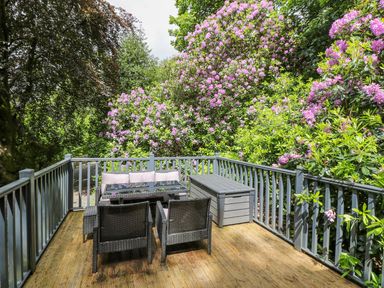 An outdoor deck with wicker furniture a table and a storage bench surrounded by blooming plants at Serenity Skiptory Howe 10 White Cross Bay near Windermere