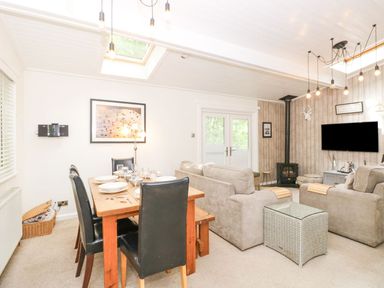 A living and dining area with a wooden dining table set with plates and glasses and beige sofas around a TV mounted on the wall at Serenity Skiptory Howe 10 White Cross Bay near Windermere