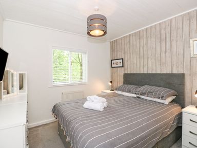 A bedroom with a double bed with striped bedding two bedside tables with lamps a window with blinds a white dresser with a mirror and a ceiling light at Serenity Skiptory Howe 10 White Cross Bay near Windermere