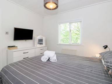 A bedroom with a bed covered in striped bedding a dresser with a mirror and a wall mounted television at Serenity Skiptory Howe 10 White Cross Bay near Windermere