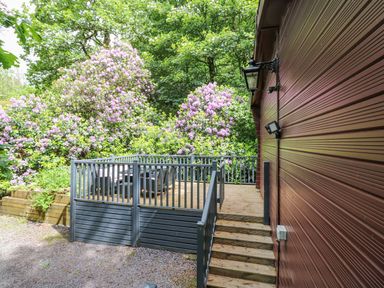 A wooden deck with outdoor furniture next to a red building surrounded by flowering bushes at Serenity Skiptory Howe 10 White Cross Bay near Windermere