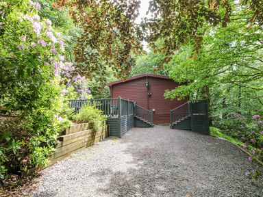 A gravel driveway leading to a brown wooden cabin with black railings and surrounded by green trees and flowering plants at Serenity Skiptory Howe 10 White Cross Bay near Windermere
