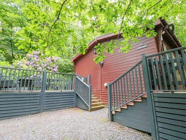 A red wooden lodge with black railings and stairs surrounded by green trees and flowering bushes at Serenity Skiptory Howe 10 White Cross Bay near Windermere