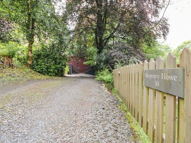 A gravel driveway leading to a wooden gate surrounded by trees and a wooden fence with a sign reading Skiptory Howe 10 at Serenity Skiptory Howe 10 White Cross Bay near Windermere