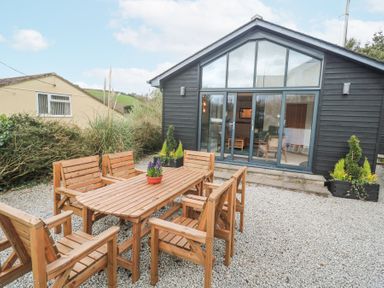 An outdoor patio with a wooden table and six chairs in front of a black wooden building with large glass doors at Chy Benyngoth in Hayle
