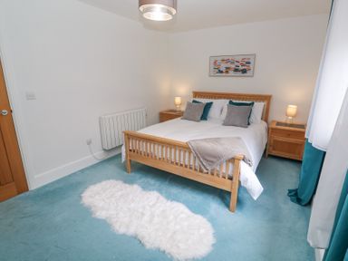 A bedroom with a wooden double bed with pillows and a throw a white rug on a blue carpet two wooden bedside tables with lamps and a painting on the wall at Chy Benyngoth in Hayle