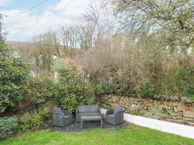 An outdoor seating area with chairs a loveseat and a table surrounded by greenery and trees at Chy Benyngoth in Hayle