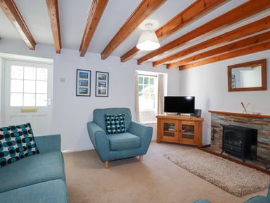 A living room with wooden beams on the ceiling blue armchairs a wooden TV stand with a television and a fireplace at Pennine in Veryan