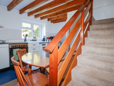 A kitchen with wooden dining table and chairs next to carpeted stairs with wooden railing at Pennine in Veryan