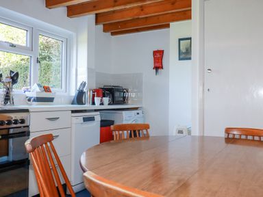 A kitchen with wooden table and chairs a window appliances and a fire blanket on wall at Pennine in Veryan