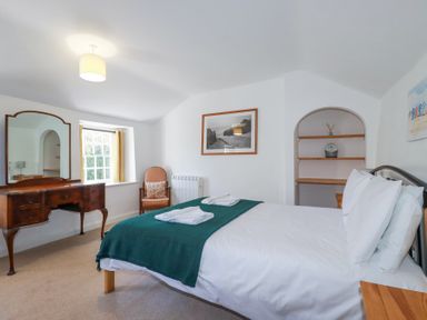 A bedroom with a bed with white linens and green throw blanket a wooden dresser with mirror an armchair and shelves at Pennine in Veryan