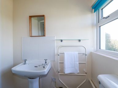 A bathroom with a white pedestal sink a small wall mirror a towel rack with a white towel and a window at Pennine in Veryan