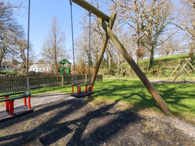A playground with two wooden and red toddler swings and a fenced area with a green playhouse at Pennine in Veryan