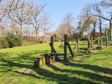 A playground with wooden climbing structures and a picnic bench on a grassy area with trees and a white building in the background at Pennine in Veryan