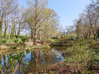 A pond surrounded by trees and bushes with a wooden fence and grassy hills in the background at Pennine in Veryan