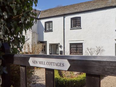 A white cottage with black-framed windows and an open door behind a gate with New Mill Cottages sign at 2 New Mill Cottages in North Bovey
