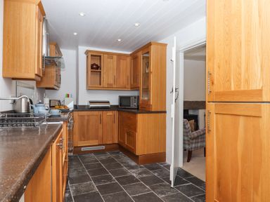 A kitchen with wooden cabinets black countertops a microwave and a door leading to a room with a chair at 2 New Mill Cottages in North Bovey