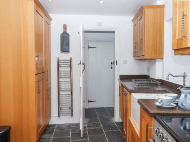 A kitchen with wooden cabinets a white door a black tile floor and blue teacups on the countertop at 2 New Mill Cottages in North Bovey
