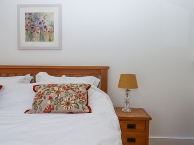 A bedroom with a wooden bed frame white bedding floral pillow wooden nightstand and a lamp at 2 New Mill Cottages in North Bovey