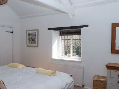 A bedroom with a bed, two rolled yellow towels, a window with blinds, a framed picture, a wicker basket, and a dresser at 2 New Mill Cottages in North Bovey