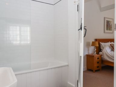 A bathroom with a bathtub and white tiled walls next to a bedroom with a wooden nightstand and lamp at 2 New Mill Cottages in North Bovey