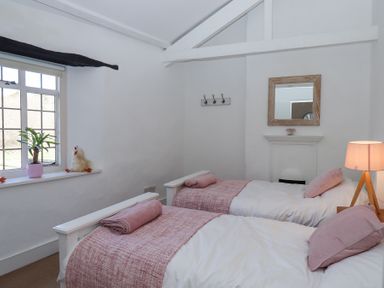 A bedroom with two single beds with pink pillows and blankets a window with a plant and toy a lamp and a wall mirror at 2 New Mill Cottages in North Bovey