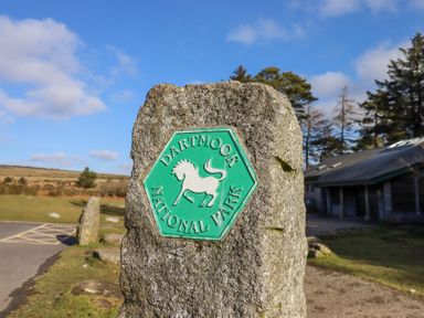 A stone sign with Dartmoor National Park emblem near a parking area with trees and a wooden building in the background at 2 New Mill Cottages in North Bovey