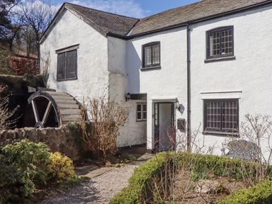 A white building with black window frames and a water wheel surrounded by bushes and a gravel path at 2 New Mill Cottages in North Bovey