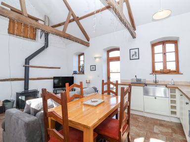 A kitchen and dining area with wooden beams on the ceiling a wooden dining table with chairs a gray sofa a black stove television and windows at The Yarde Annexe in Pen Elm near Taunton
