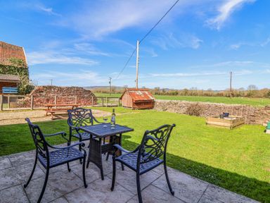 A patio with four metal chairs and a table with a bottle and glasses overlooking a grassy yard with a wooden shed and picnic bench at The Yarde Annexe in Pen Elm near Taunton