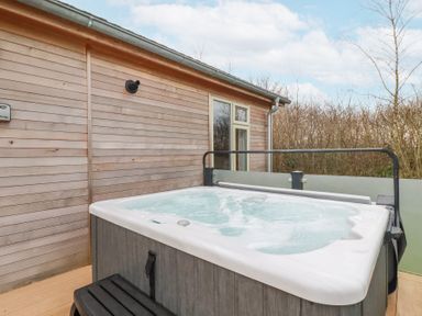 An outdoor hot tub on a wooden deck next to a wooden house wall with trees in the background at 6 Meadow Retreat in Dobwalls