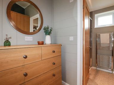 A wooden dresser with knobs and a round mirror above it next to a doorway leading to a bathroom with a towel on a radiator and patterned floor at 6 Meadow Retreat Dobwalls