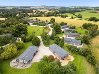 An aerial view of multiple small houses along curved gravel paths surrounded by green fields and trees at 6 Meadow Retreat in Dobwalls