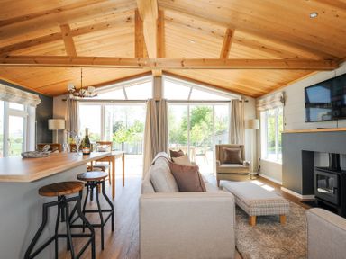 A living room with beige sofa armchair plaid ottoman wood stove and kitchen bar stools at 17 Meadow Retreat in Dobwalls