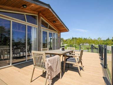 An outdoor deck with wooden table and chairs by a building with large windows at 17 Meadow Retreat in Dobwalls