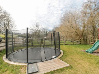 An outdoor fenced grassy area with a round trampoline and a green slide at Bell tent 3 in Camerton near Timsbury