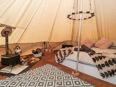 Interior of a tent with a bed, wood stove, firewood, kettle, cushions, blankets, and a hanging candle holder at Bell tent 4 in Camerton near Timsbury