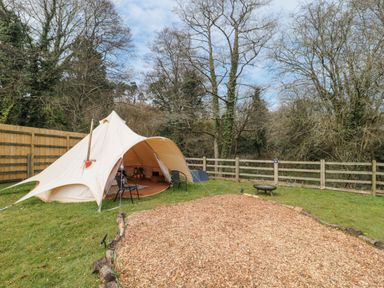 A bell tent with chairs on grass near a fire pit and wood chip area surrounded by trees and fencing at Bell tent 4 in Camerton near Timsbury