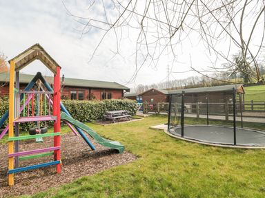 A playground with a slide swing and trampoline next to wooden buildings at Bell tent 4 in Camerton near Timsbury
