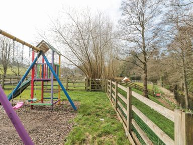 A children's playground with swings and a slide near a wooden fence and grassy area at Bell tent 4 in Camerton near Timsbury