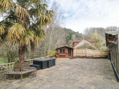 An outdoor patio area with a palm tree surrounded by a wooden bench a black table with stools a wooden shed in the background and a house behind a wooden fence at Bell tent 4 Camerton near Timsbury