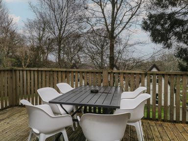 A wooden outdoor deck with a black table and six white chairs surrounded by trees at Valley Lodge 38 in St Ann's Chapel Cornwall