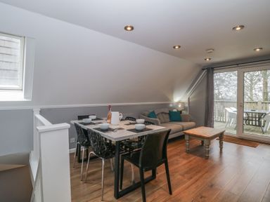 An open plan living and dining area with a dining table set with bowls and utensils a sofa with cushions a wooden coffee table and sliding glass doors opening to a balcony at Valley Lodge 38 in St Ann's Chapel Cornwall