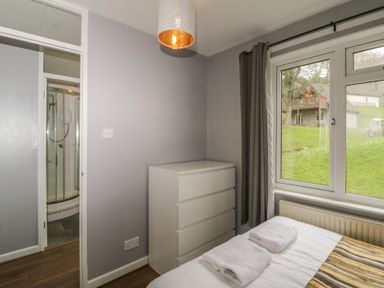 A bedroom with a bed with towels on it a white chest of drawers a window with curtains and a door leading to a bathroom with a shower at Valley Lodge 38 St Ann's Chapel Cornwall
